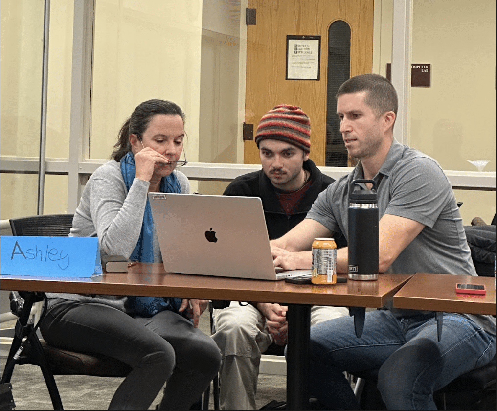 Three people are focused on a laptop in a classroom setting. A blue card with "Ashley" is visible on the table.