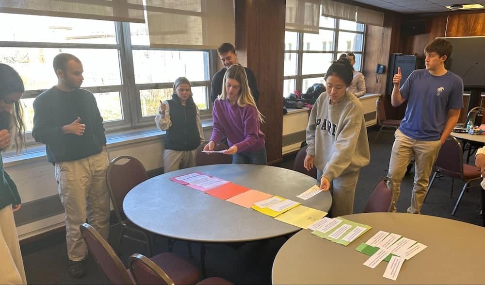 A group of students engage in a card-sorting activity around tables in a well-lit room, participating in a collaborative exercise.