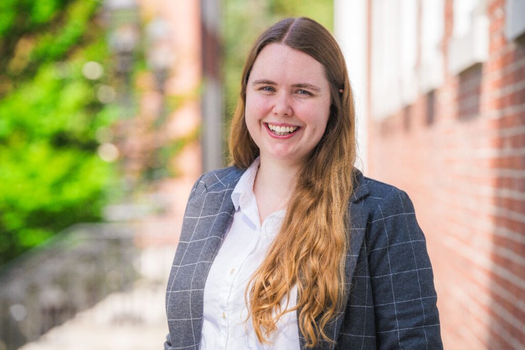 Photo of Dr. Emma Klugman, Program Director of Data Science, smiling outdoors in a white shirt and a gray checkered blazer.